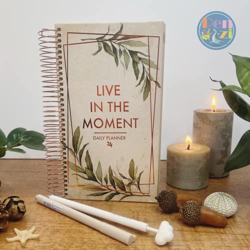 دفتر برنامه‌ریزی روزانه فنری با جلد سخت و طرح برگ‌های گیاه، با عنوان انگلیسی «LIVE IN THE MOMENT»، کنار شمع و خودکار روی میز چوبی