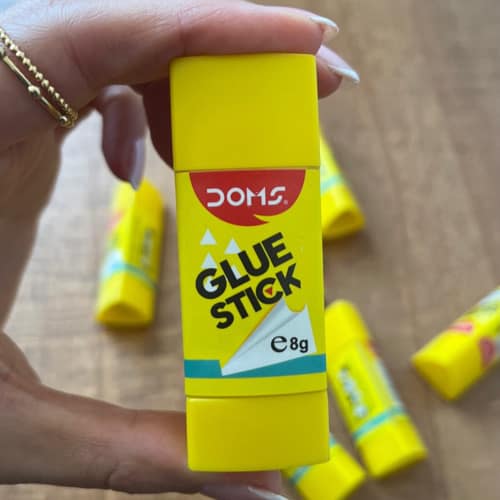 چسب استیکی زردرنگ دامس با برچسب انگلیسی GLUE STICK، در دست نگه داشته شده و چند چسب مشابه در پس‌زمینه دیده می‌شود.