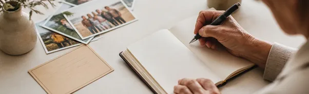 دست سالمندی در حال نوشتن در دفتر خاطراتی باز، کنار عکس‌های خانوادگی و فنجان چای روی میز