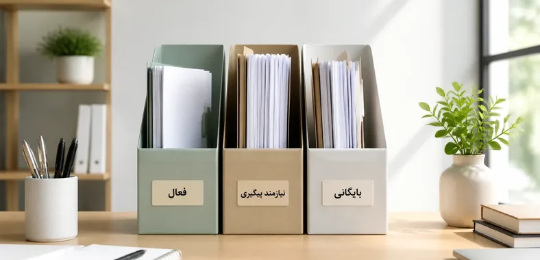 سه جعبه بایگانی رومیزی حاوی کاغذها، با برچسب‌های «فعال»، «نیازمند پیگیری» و «بایگانی»، روی میز کار اداری